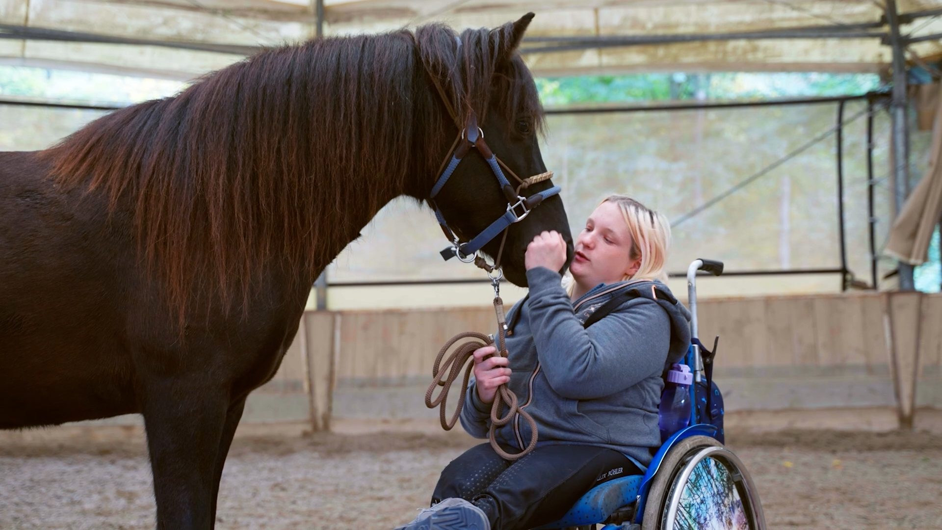 Lisa Vas, eine junge Frau mit blondem Haare im Rollstuhl, streichelt ihr großes, dunkelbraunes Pferd