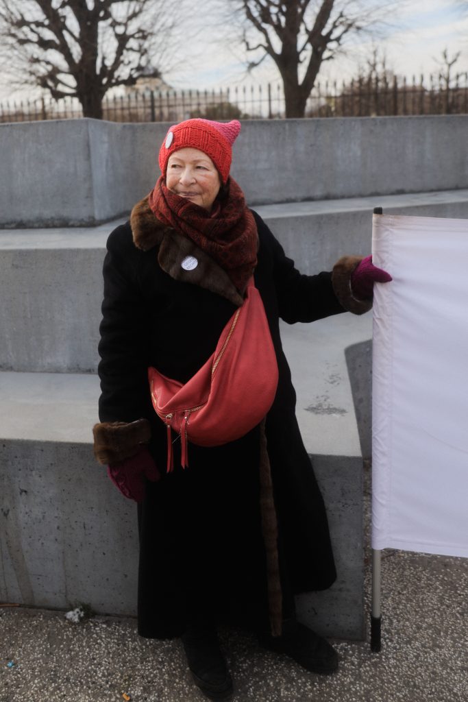Oma gegen rechts. Sie trägt einen schwarzen Mantel eine rote Umhängetasche und eine rote Mütze