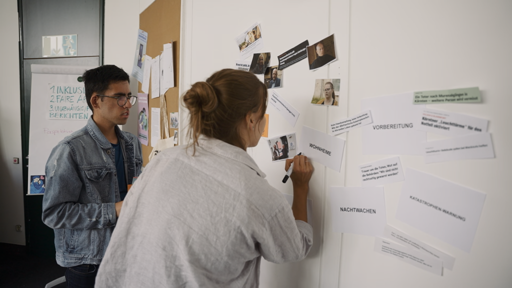 Clara und Artin stehen an einer Wand, auf der Fotos und ausgedruckte Texte hängen.