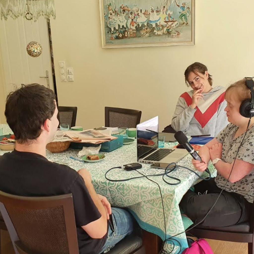 Drei Personen sitzen in einem Wohnzimmer. Die Frau in der Mitte nimmt mit einem Mikrofon in der Hand und Kopfhörern auf.