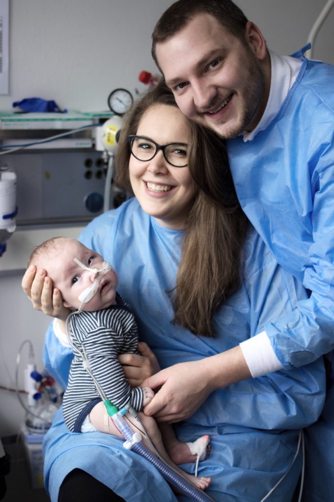 Lisa und ihr Mann tragen Krankenhaus-Kittel und lächeln in die Kamera. Sie halten ihr Neugeborenes.