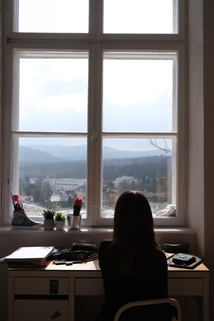 Ein Mädchen sitzt mit dem Rücken zur Kamera vor einem Fenster und blickt nach draussen.