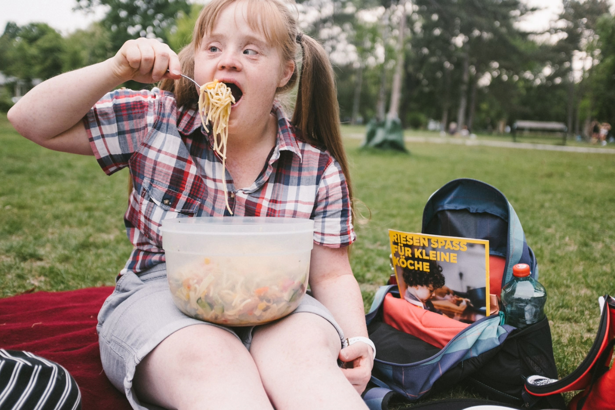 Hanna Gugler sitzt auf einer Picknickdecke. Sie isst einen Nudelsalat. Ihre Haare sind blond und zu zwei Zöpfen gebunden.