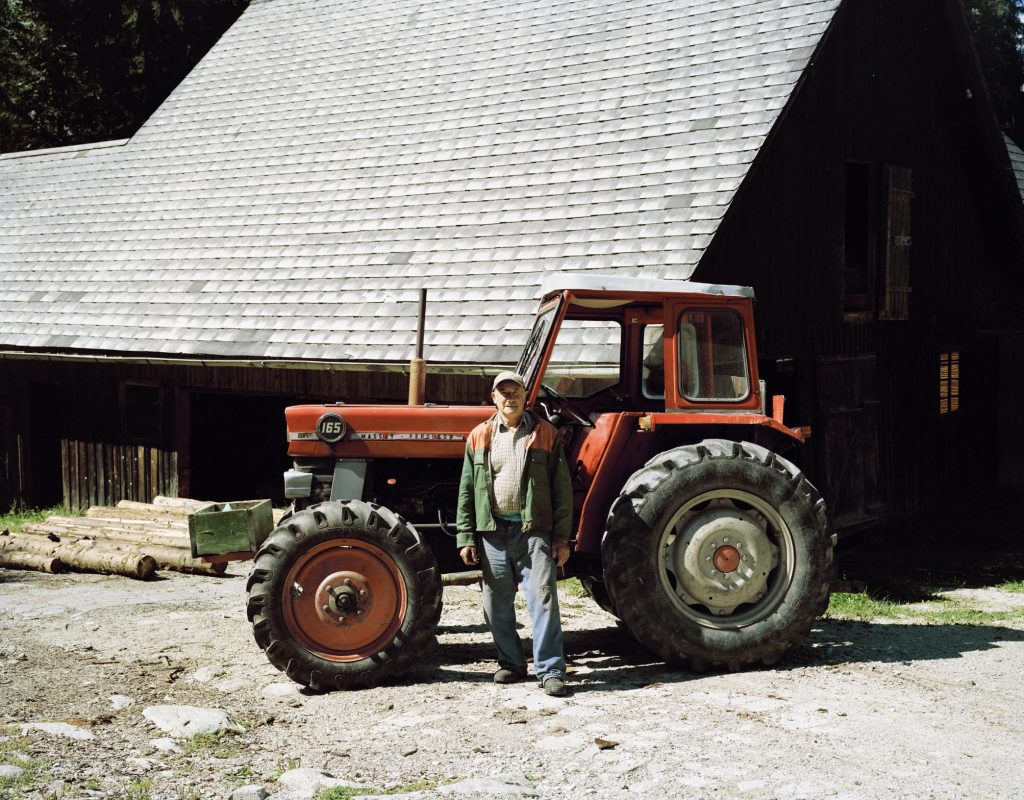 Ein Mann steht vor einem roten Traktor. Er trägt eine Mütze und schaut mit ernsten Blick in die Kamera. Im Hintergrund ist ein Haus.