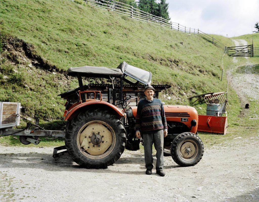 Ein älterer Mann steht vor einem roten Traktor. Er trägt eine Mütze und schaut in die Kamera. Im Hintergrund sieht man einen Hügel und Holzzaun.