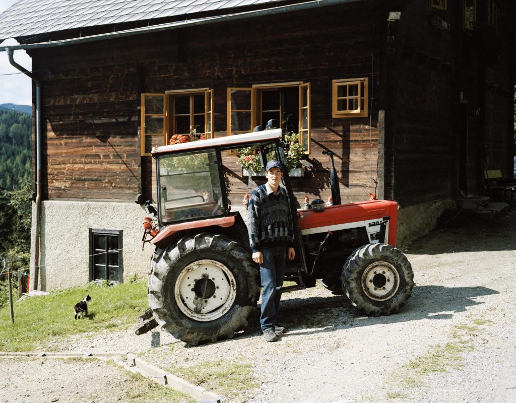 Ein junger Mann steht vor einem roten Traktor. Er schaut mit ernsten Blick in die Kamera. Links von ihm ist eine Katze. Im Hintergrund ist ein Bauernhaus.