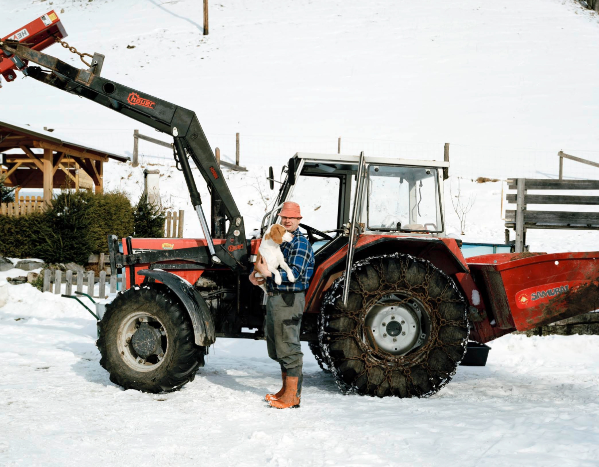 Ein älterer Mann steht vor einem Traktor. Er trägt eine rote Mütze. In den Händen hält er einen Hund. Auf den Boden liegt Schnee.