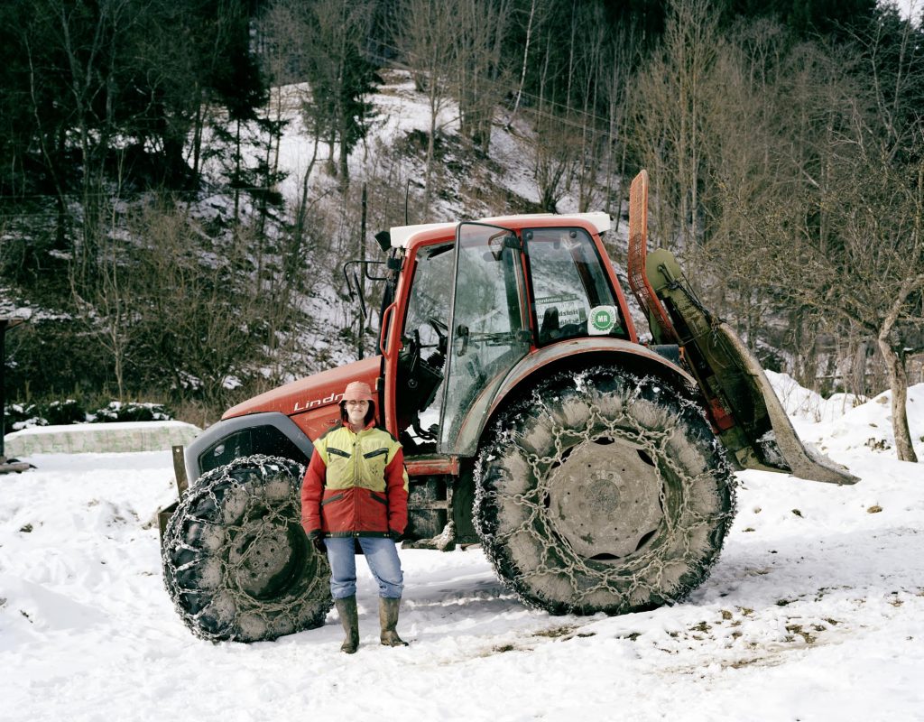 Eine Frau steht vor einem roten Traktor und lächelt in die Kamera. Sie trägt eine rote Mütze und warme Kleidung. Schnee liegt auf den Boden.