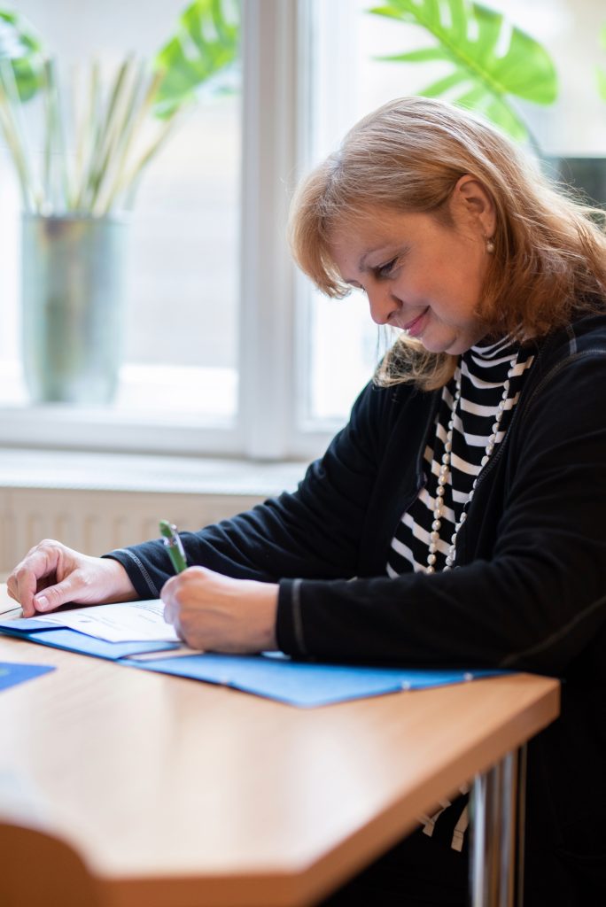 Petra Smutny sitz an einem Tisch. Sie schreibt in den Unterlagen. Sie trägt blonde Haare und blickt auf ihre Arbeitsfläche.