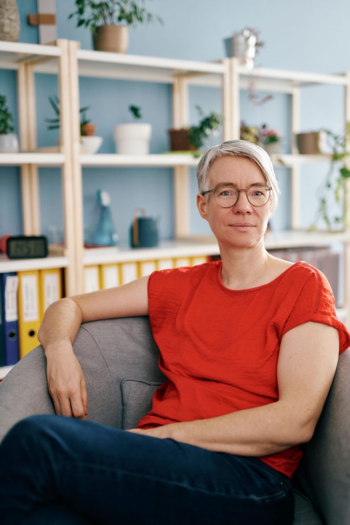 Ein Portrait von Lisa Udl. Sie sitzt auf einem Stuhl, trägt eine Brille und ein rotes T-Shirt. Sie schaut in die Kamera.