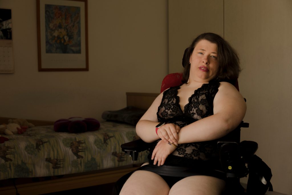 Leonie Schüler sitzt in ihrem Rollstuhl. Sie schaut mit ernsten Blick in die Kamera. Sie trägt einen schwarzen Spitzen-Body und pinken Lippenstift.