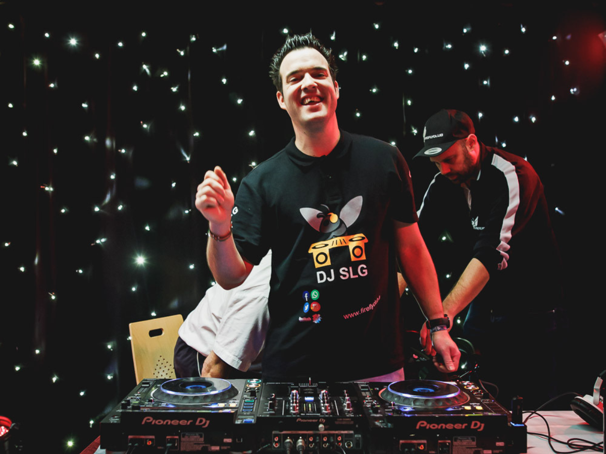 Das Foto zeigt Sebastian Gruber bei seiner Arbeit als DJ. Er steht vor einem Mischpult und lacht in die Kamera. Im Hintergrund sind farbige Lichter.