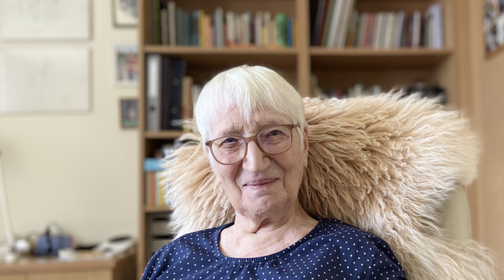 Traude Veran sitzt in einem Lehnsessel mit Schaffell und blickt in die Kamera. Sie hat kurze weiße Haare und trägt eine Brille, im Hintergrund ein volles Bücherregal.