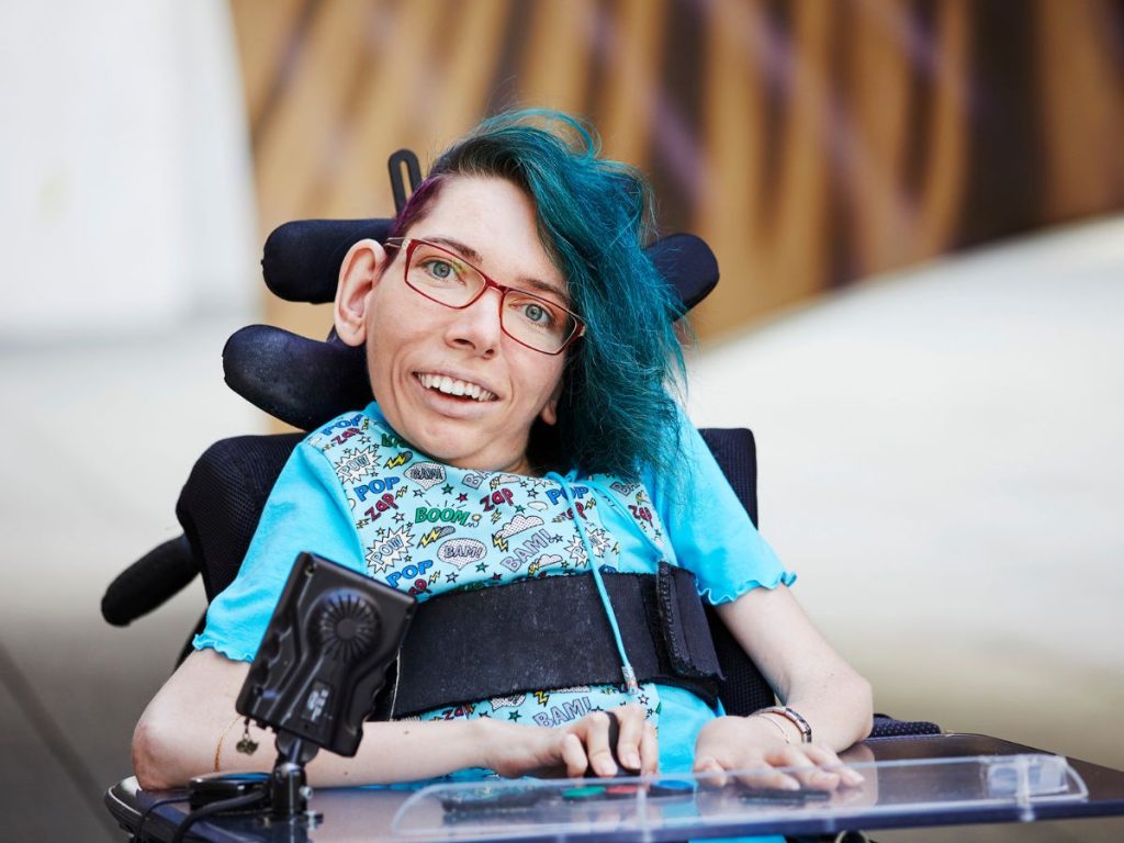 Melanie Eilert schaut mit freundlichem Blick in die Kamera. Sie hat blaue Haare, trägt eine Brille und sitzt in einem Rollstuhl.