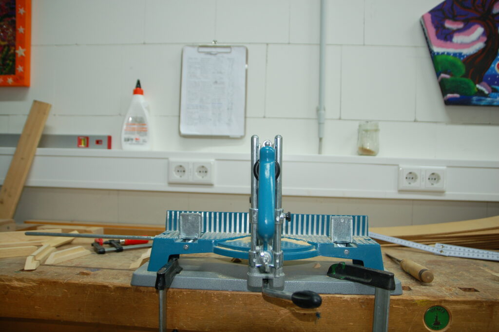 Ein Arbeitstisch in einer Werkstatt, auf dem ein blaues und silbernes Werkzeug für Holzbearbeitung (eine Fräsvorrichtung) steht. Im Hintergrund hängt ein Clipboard mit Papieren an der Wand. Daneben sind eine Flasche Holzleim und einige Holzlatten zu sehen.