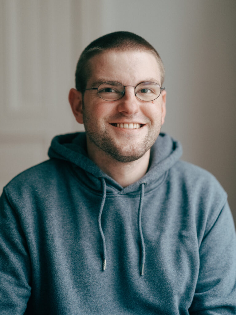 Profilfoto von Richard. Er ist ein junger Mann mit kurzen Haaren und Brille, der einen Blauen Hoodie trägt und in die Kamera lächelt.