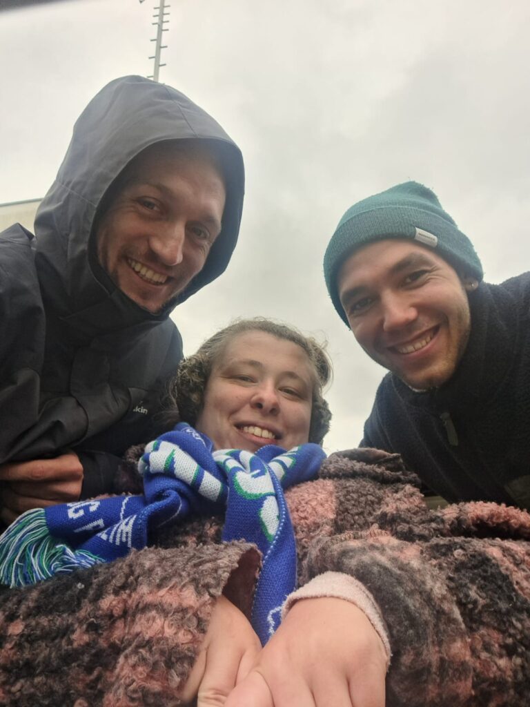 Ein Selfie von drei Personen vor grauem Himmel. Leonie trägt einen blau-weißen Schal und ist in der Mitte zu sehen, links und rechts von ihr ist jeweils ein junger Mann mit Mütze bzw. Kapuze. Sie lächeln alle drei in die Kamera.