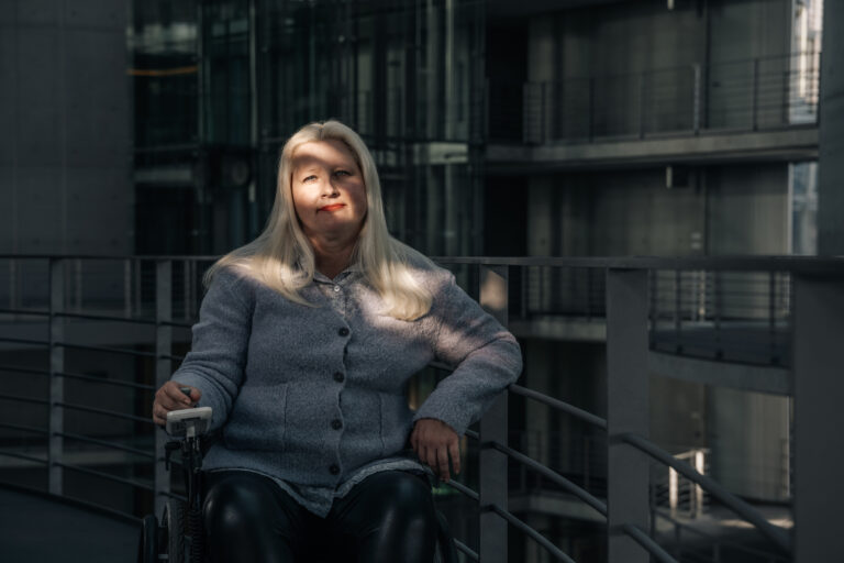 Eine Frau im Rollstuhl blickt in die Kamera. Sie hat lange blonde Haare.