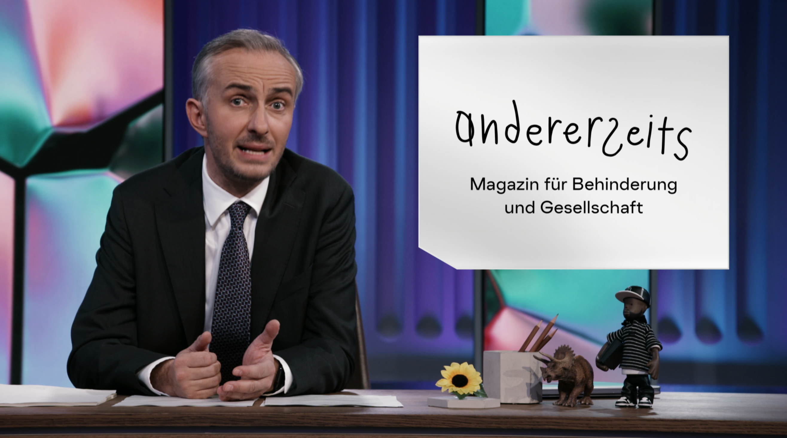 Das Bild zeigt Jan Böhmermann, einen bekannten Moderator, der hinter einem Tisch sitzt. Er trägt einen schwarzen Anzug, ein weißes Hemd und eine gestreifte Krawatte. Rechts neben ihm steht ein Schild mit der Aufschrift „andererseits – Magazin für Behinderung und Gesellschaft“. Auf dem Tisch liegen Papiere, ein kleiner Dinosaurier, ein kleines Männchen in Baseballkleidung, eine gelbe Blume und eine Box mit Stiften. Der Hintergrund ist bunt und modern gestaltet.