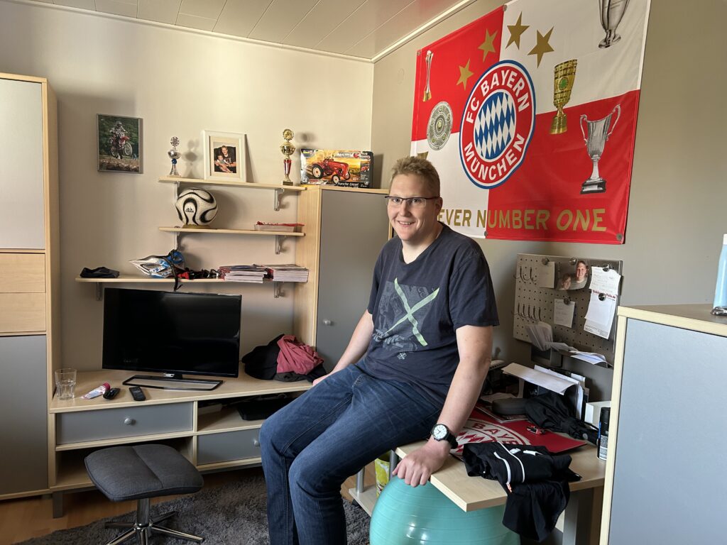 Sebastian Wirth schaut mit freundlichen Blick in die Kamera. Er hat kurze Haare und trägt eine Brille. Im Hintergrund sieht man ein Wohnzimmer mit mehreren Regalen. An der Wand hängt ein Poster mit "FC Bayern München".