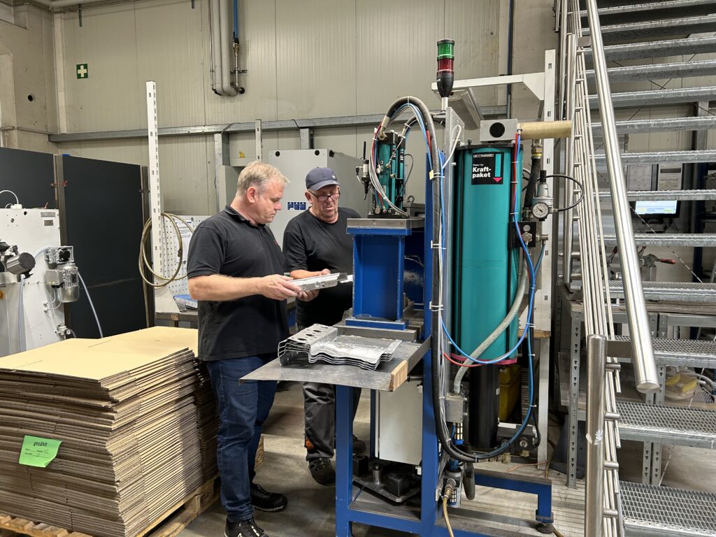 Zwei Männer stehen in einer Werkstatt. Sie arbeiten an einer blauen Maschine. Ein Mann hält ein Metall-Stück in den Händen.