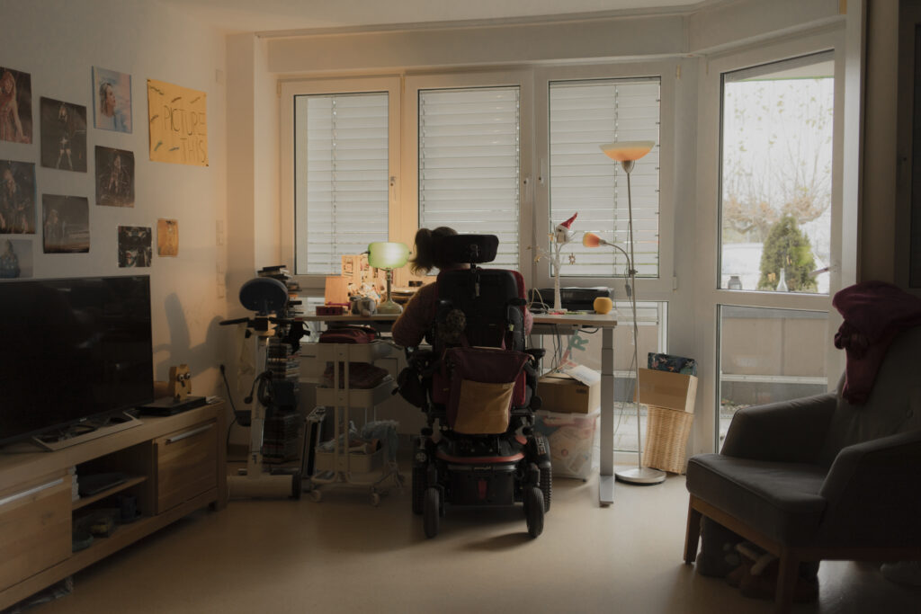 Ein gemütlich eingerichtetes Wohnzimmer. Vor dem Fenster steht ein Schreibtisch. Davor sitzt Leonie mit dem Rücken zur Kamera.