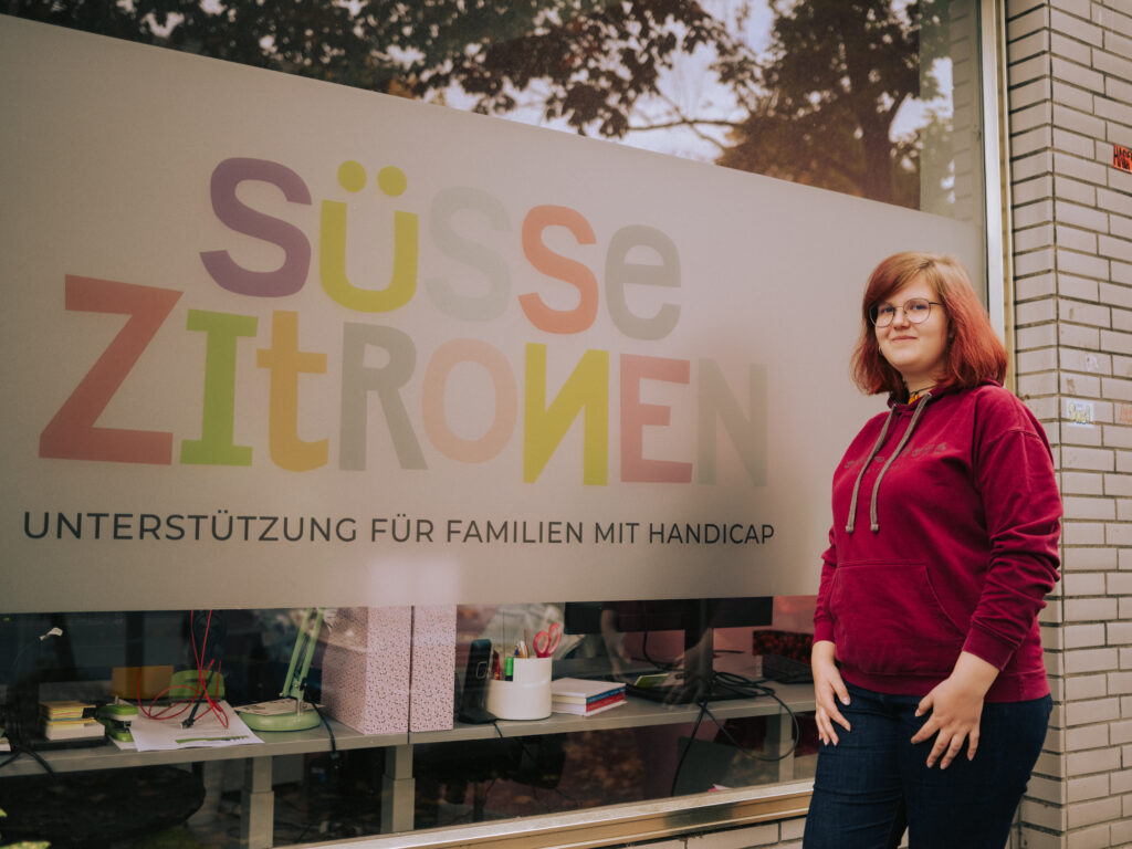 Lisa K. steht vor einem Gebäude. Sie hat kurze Haare und einen roten Pullover an.