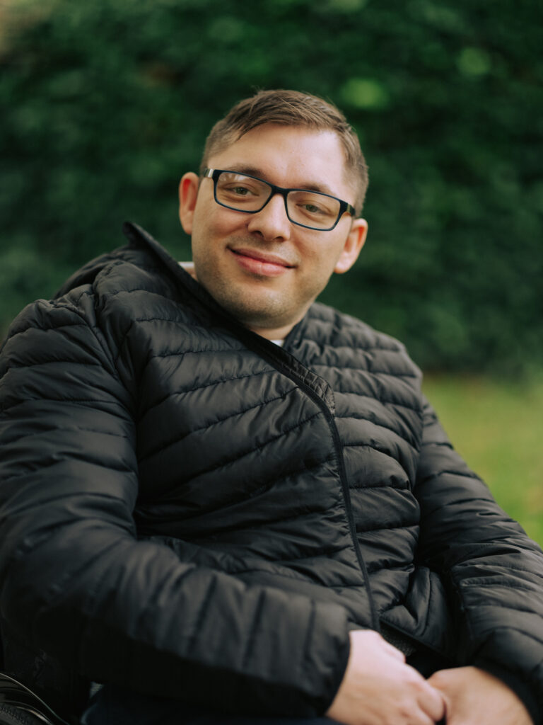 Lulzim Lushtaku schaut mit freundlichen Blick in die Kamera. Er trägt eine schwarze Winterjacke und trägt eine Brille.