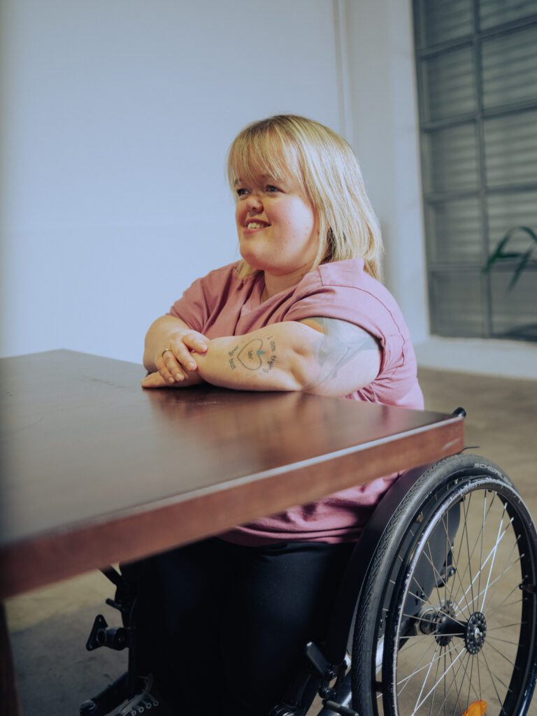 Eine junge Frau mit kurzen blonden Haaren und einem pinken Oberteil sitzt an einem Tisch. Sie verwendet einen Rollstuhl.