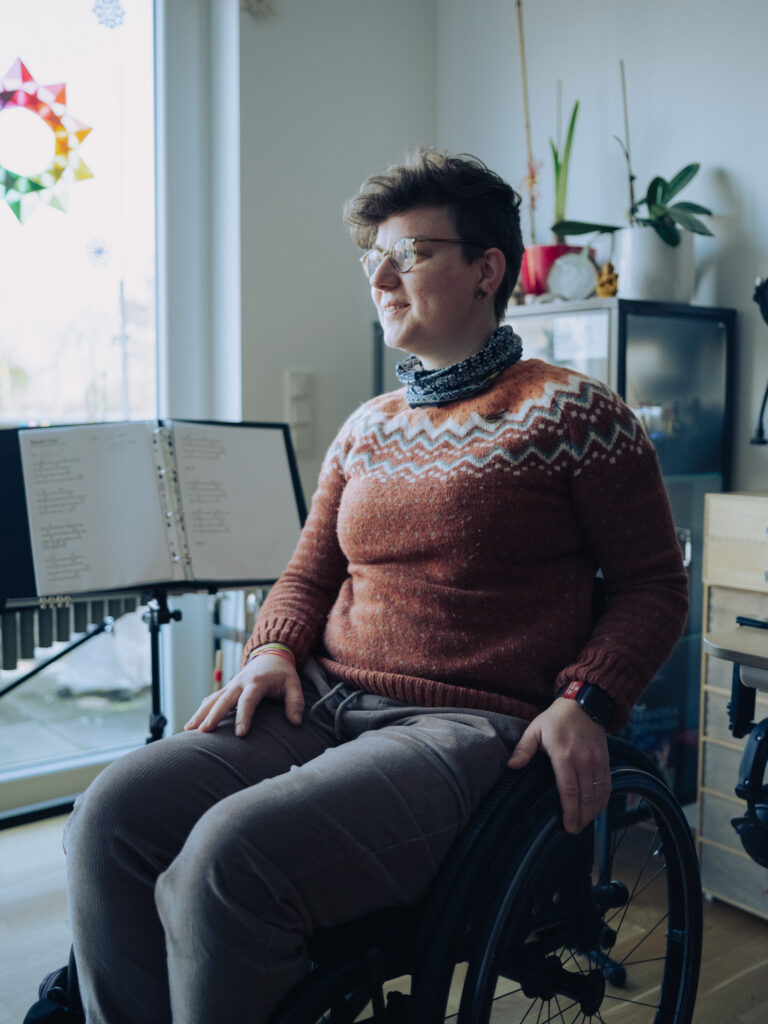 Eine Frau mit kurzen braunen Haaren, Brille, einem orangen Pullover, einem schwarz-weißen Schal und einer grauen Hose sitzt im Rollstuhl. Sie befindet sich in einem Zimmer, im Hintergrund sieht man eine aufgeschlagene Musikmappe. Sie blickt lächelnd an der Kamera vorbei.