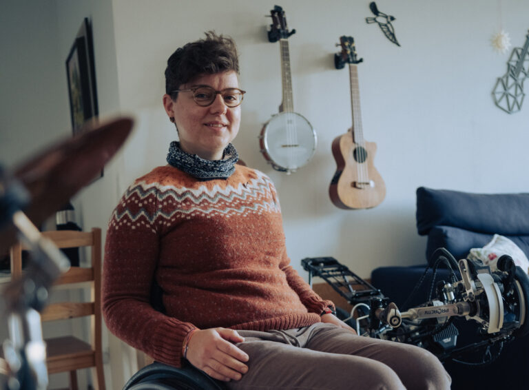 Eine Frau mit kurzen braunen Haaren, Brille, einem orangen Pullover, einem schwarz-weißen Schal und einer grauen Hose sitzt im Rollstuhl. Sie befindet sich in einem Zimmer, im Hintergrund sieht man eine aufgeschlagene Musikmappe. Sie blickt lächelnd an der Kamera vorbei.