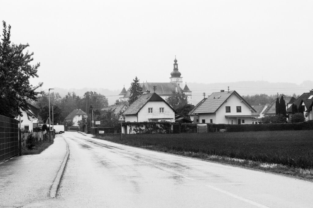 Das Foto zeigt eine schwarz-weiß Aufnahme. Eine Straße führt in ein Dorf auf der linken Seite ist ein Gehsteig, auf der rechten Seite ein Feld. Dahinter stehen Familienhäuser. Im Hintergrund sieht man einen Kirchturm und das Schloss Hartheim.