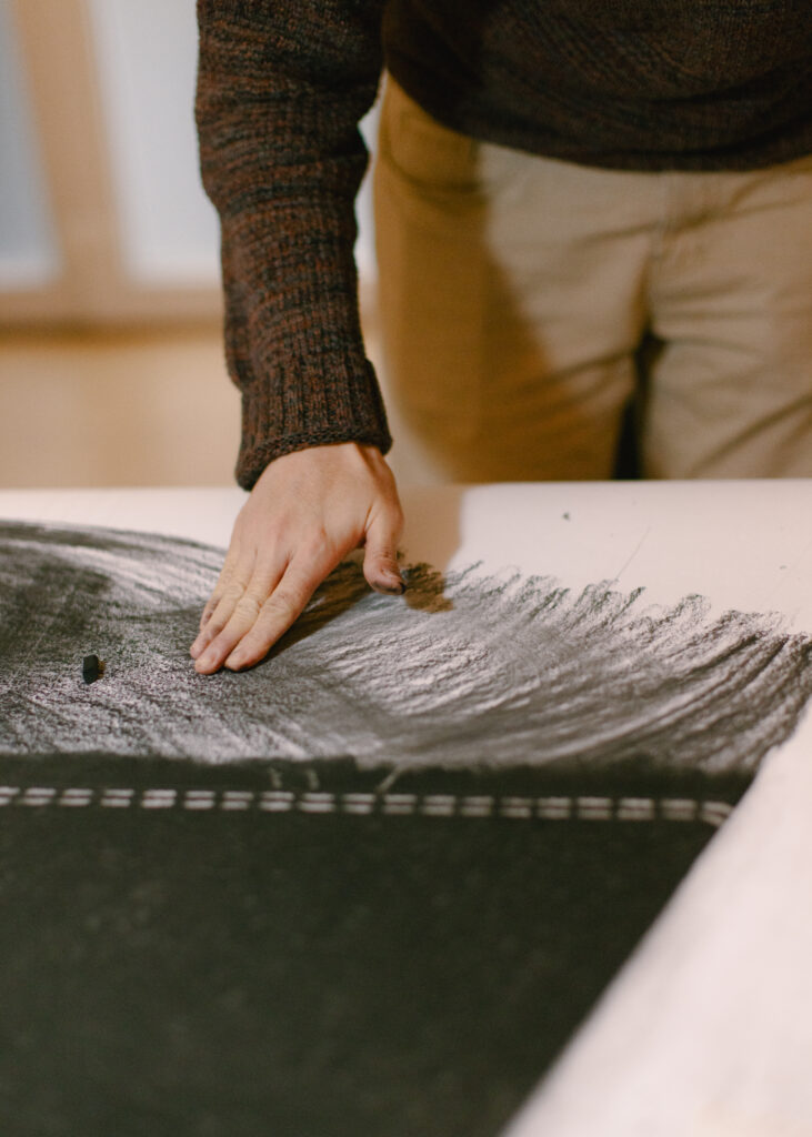 Auf dem Bild sieht man die Hand von dem Künstler Juewen Zhang. Er verwischt mit den Fingern schwarze Pastellkreide auf dem Papier.