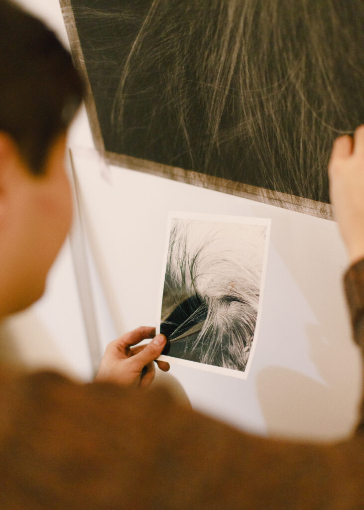 Auf dem Bild sieht man den Künstler Künstler Juewen Zhangn von hinten. Er hält eine Zeichnung in der linken Hand, auf der fein gezeichnete Haare zu sehen sind. Darüber hängt ein Bild an einer Wand, an der ebenso Haare zu sehen sind.