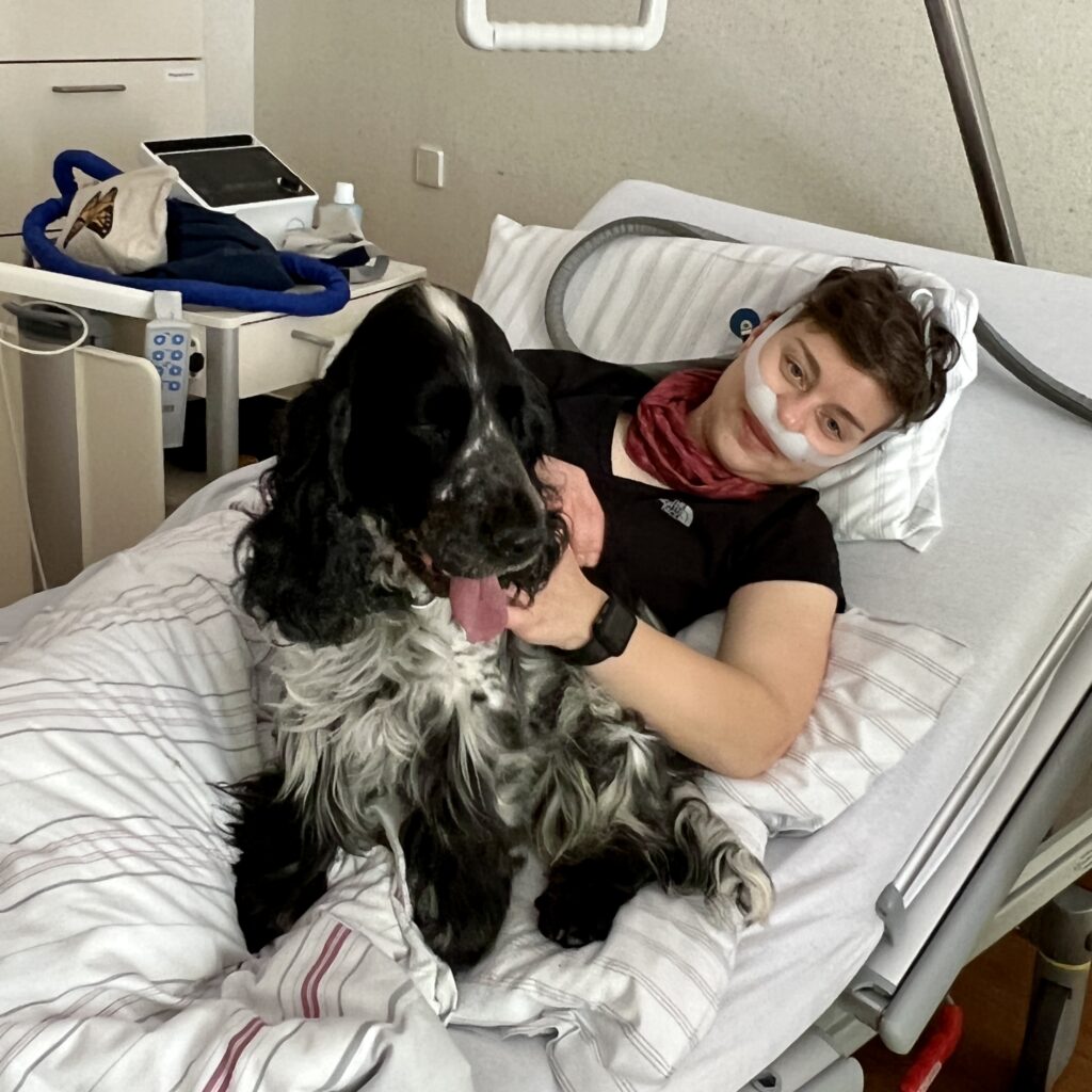 Franziska Seehausen liegt in einem Krankenhausbett. Sie sieht mit ernstem Blick in die Kamera. Sie trägt ein schwarzes T-Shirt und ein rotes Halstuch. Neben ihr sitzt ein großer schwarz-weißer Hund auf dem Bett. Im Hintergrund stehen medizinische Geräte.