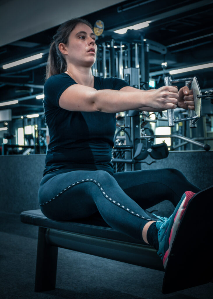 Eine junge Frau sitzt auf einer Bank im Fitnessstudio. Sie hält mit beiden Händen eine Stange und zieht daran. Sie trägt schwarze Sportkleidung und bunte Sportschuhe.