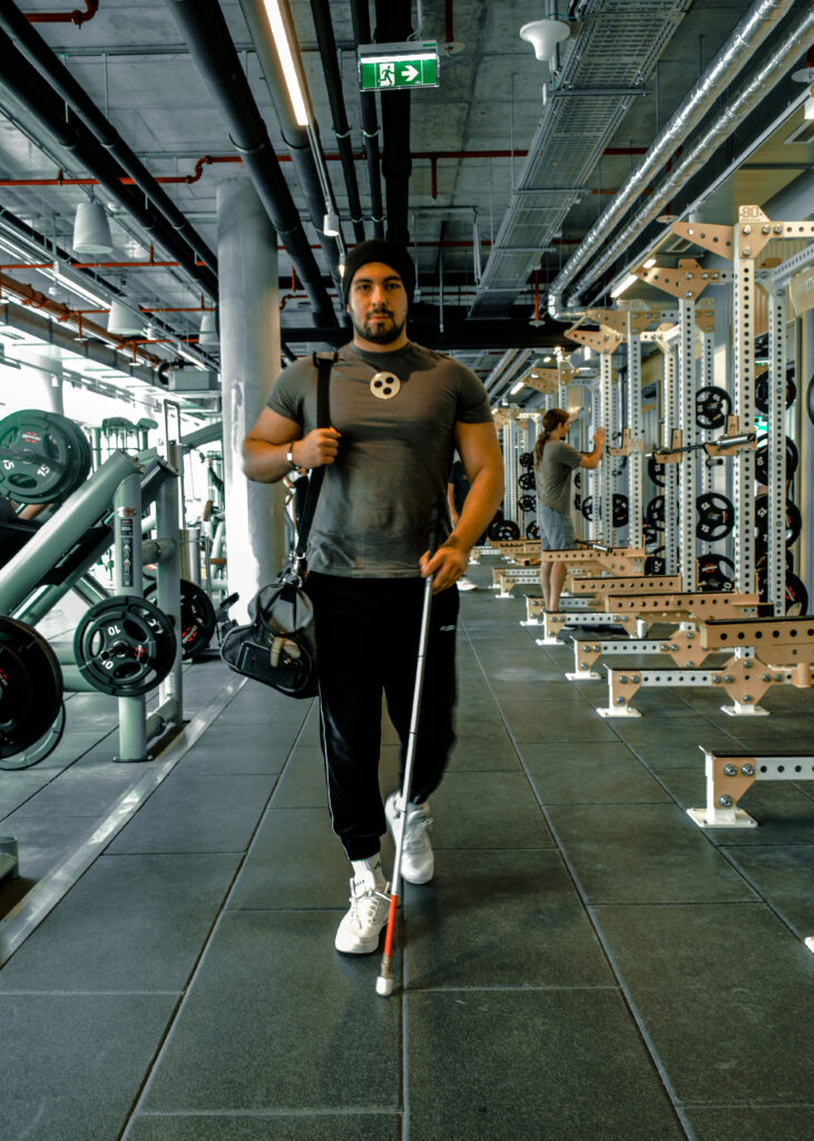 Ein junger Mann steht im Fitnessstudio. Er benutzt einen Blindenstock und ein Blindenabzeichen. Er trägt dunkle Sportkleidung. Über die rechte Schulter hängt eine Sporttasche.