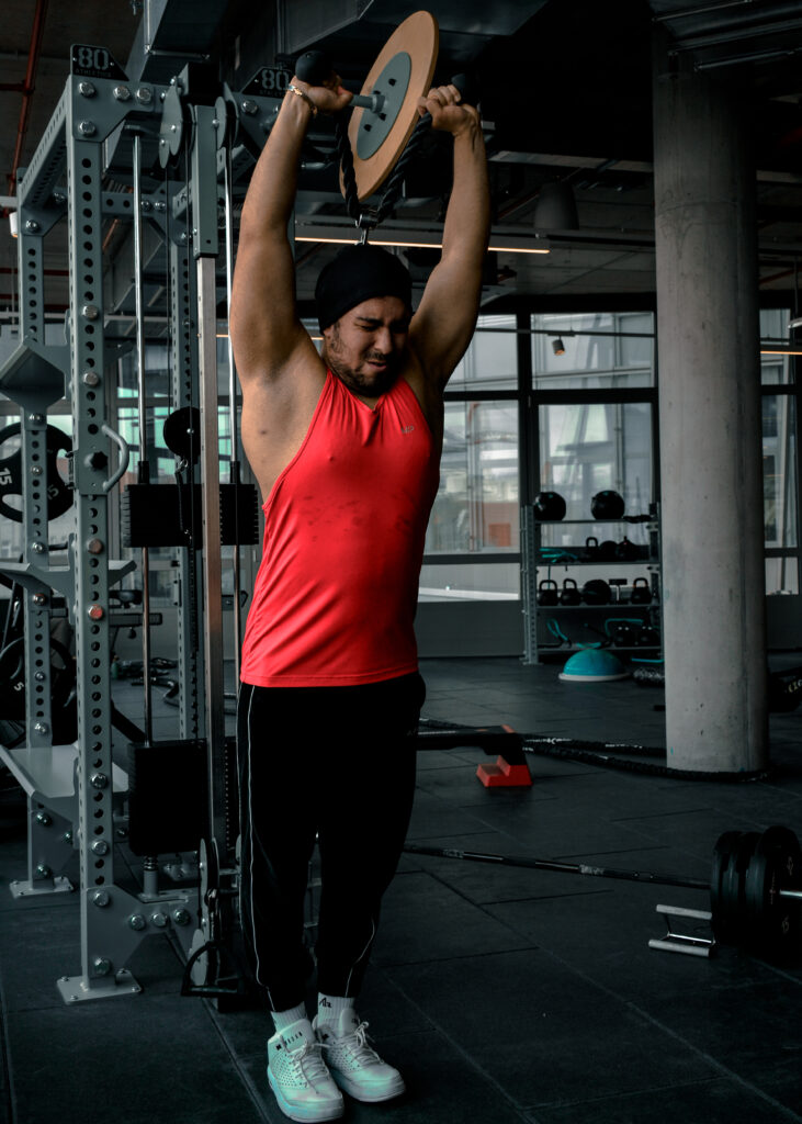 Ein junger Mann hebt in einem Fitnessstudio ein Gewicht mit beiden Händen über den Kopf. Sein Augen sind geschlossen, das Gesicht hat einen angestrengten Ausdruck.