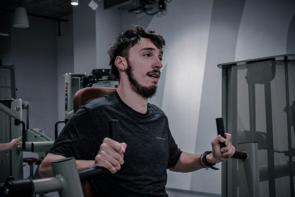 Ein junger Mann sitzt im Fitnessstudio auf einem Gerät. Er schaut mit angestrengten Blick nach vorne.