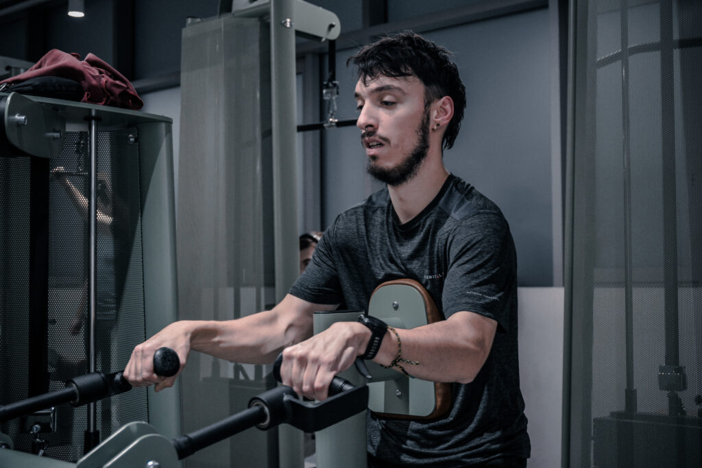 Ein junger Mann sitzt im Fitnessstudio auf einem Gerät. Seine Augen sind halb geschlossen. Sein Gesichtsausdruck ist müde.