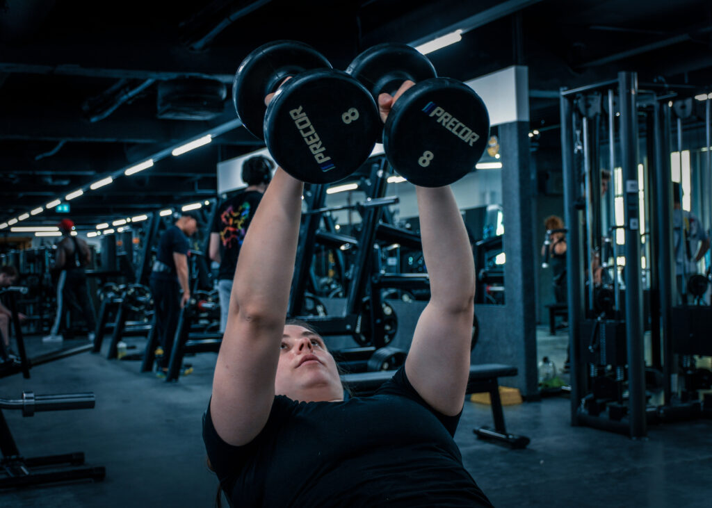 Eine junge Frau liegt auf einer Bank im Fitnessstudio. Sie hebt zwei Hanteln über ihren Kopf. Andere Menschen trainieren im Hintergrund.