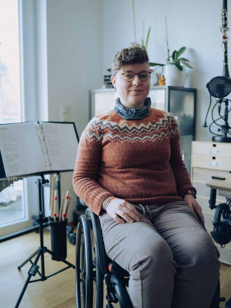 Franziska Seehausen sitzt in einem Rollstuhl und schaut mit freundlichem Gesicht in die Kamera. Sie hat kurze braune Haare, trägt einen roten Pullover und eine graue Jogging-Hose. Hinter ihr steht ein Notenständer und Regale mit Zimmerpflanzen.